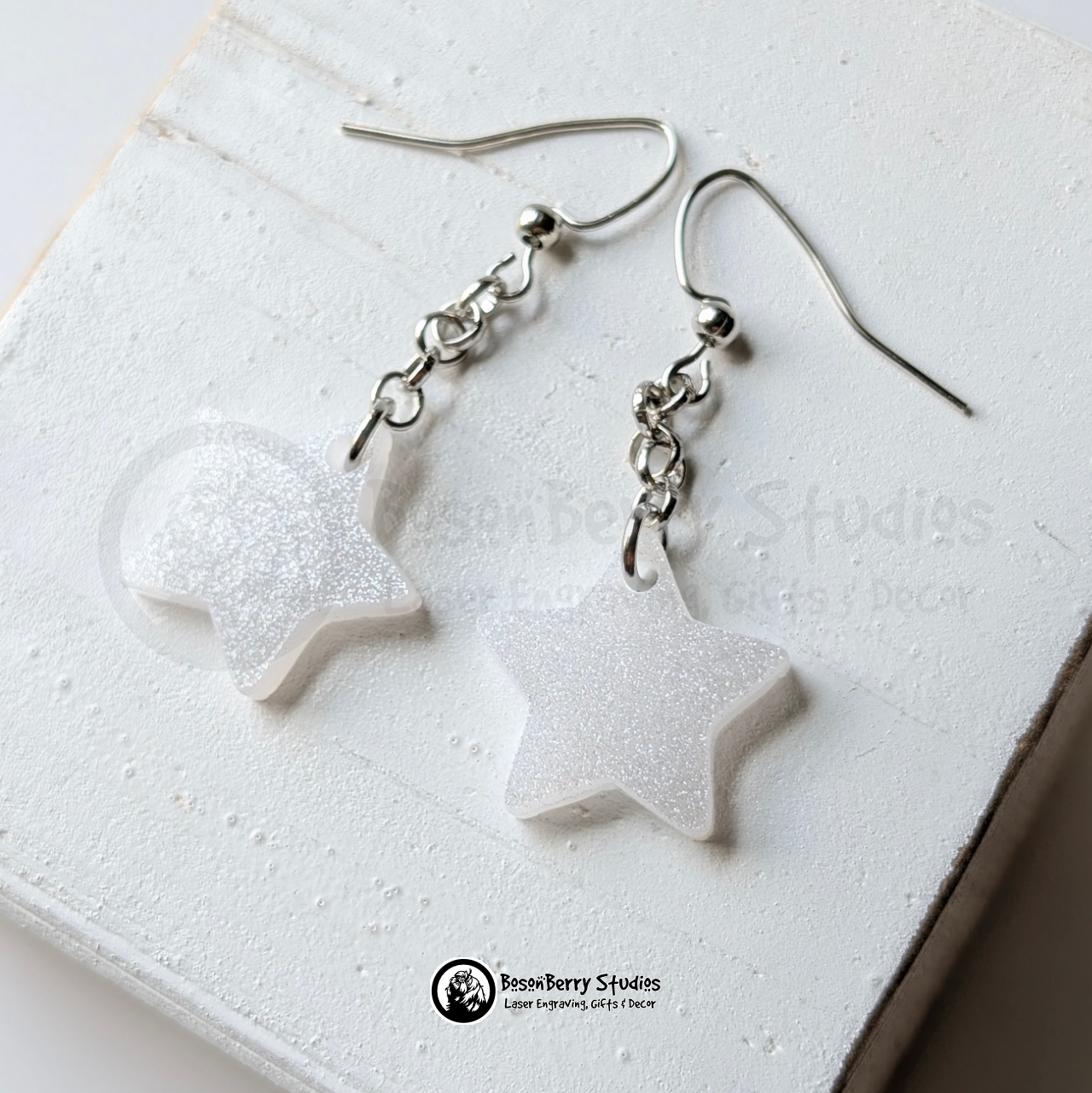 White star-shaped glitter dangle earrings on a white background with "BosonBerry Studios" branding.