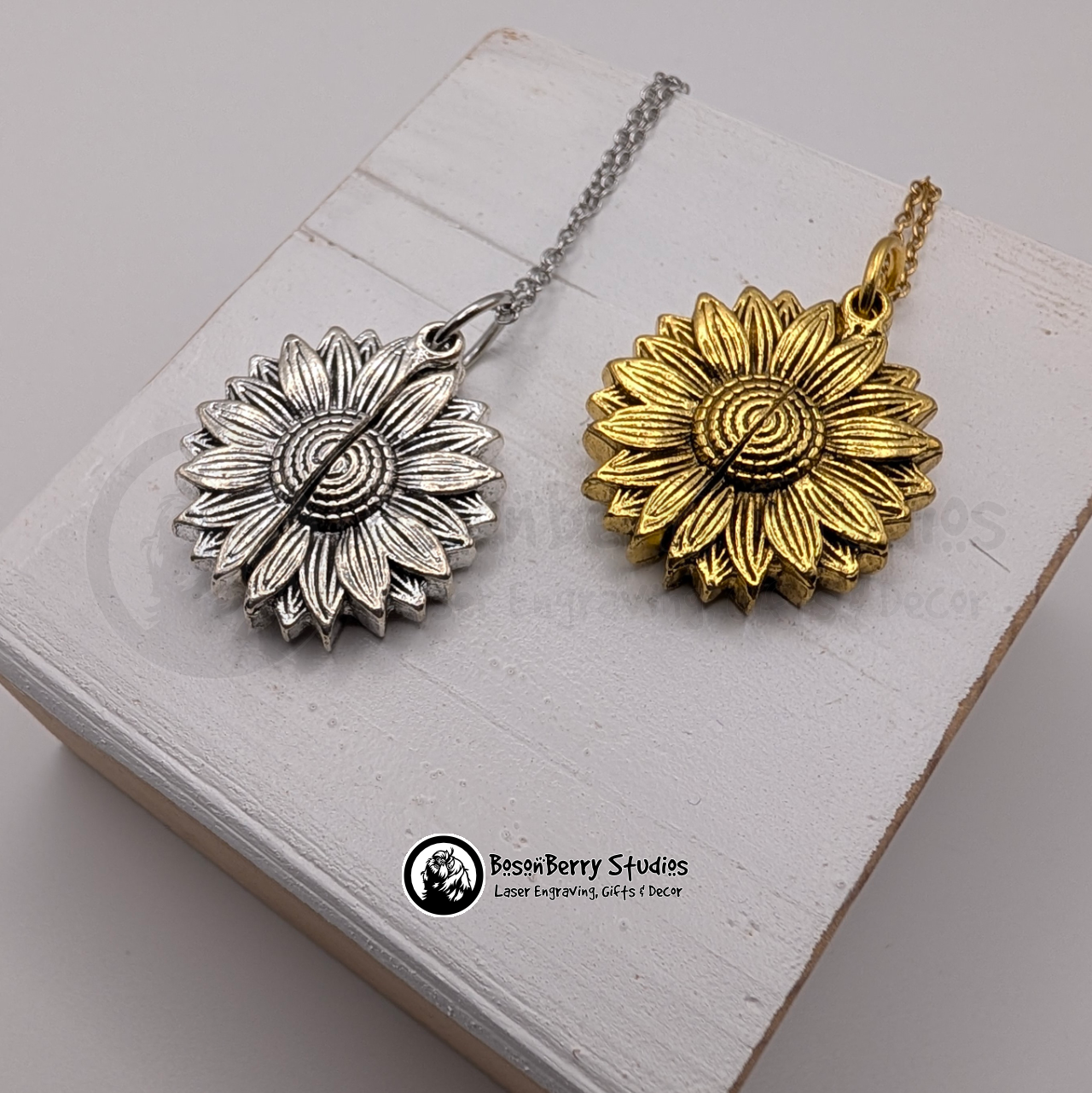 Two sunflower pendant necklaces, one gold and one silver, on a white wooden stand with 'BosonBerry Studios' branding.