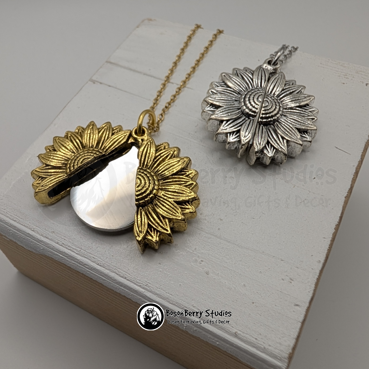 Two sunflower pendant necklaces, one gold that's open to reveal a silver disc, and one silver closed, on a white wooden stand with 'BosonBerry Studios' branding.