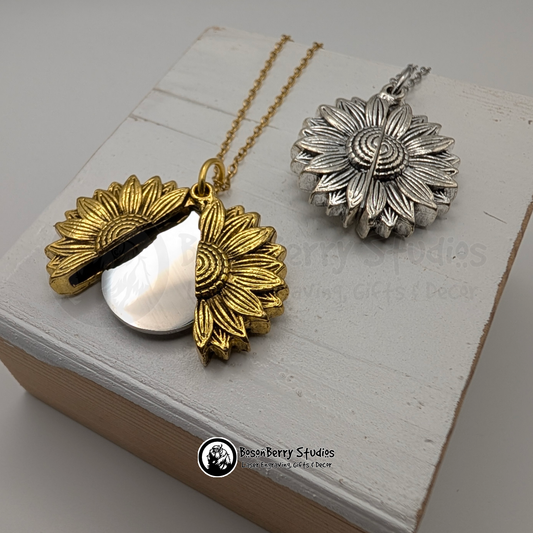 Two sunflower pendant necklaces, one gold that's open to reveal a silver disc, and one silver closed, on a white wooden stand with 'BosonBerry Studios' branding.