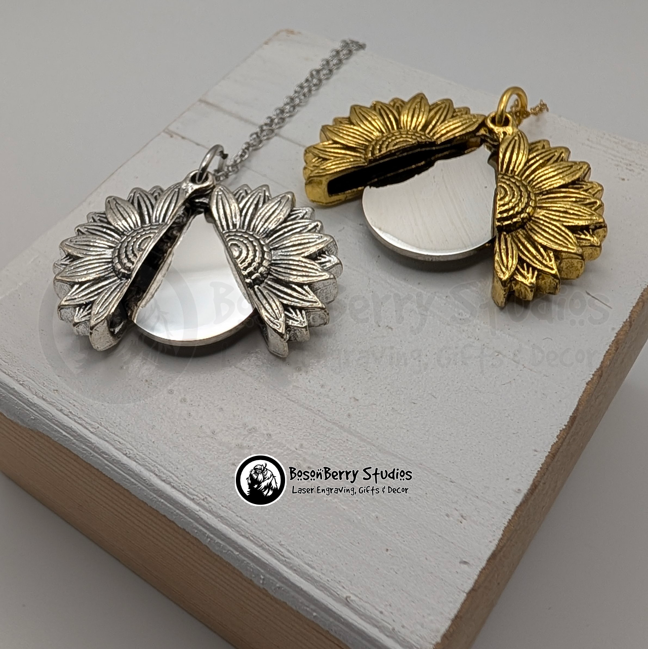 Two sunflower pendant necklaces, one gold and one silver, both open to reveal a silver disc, on a white wooden stand with 'BosonBerry Studios' branding.