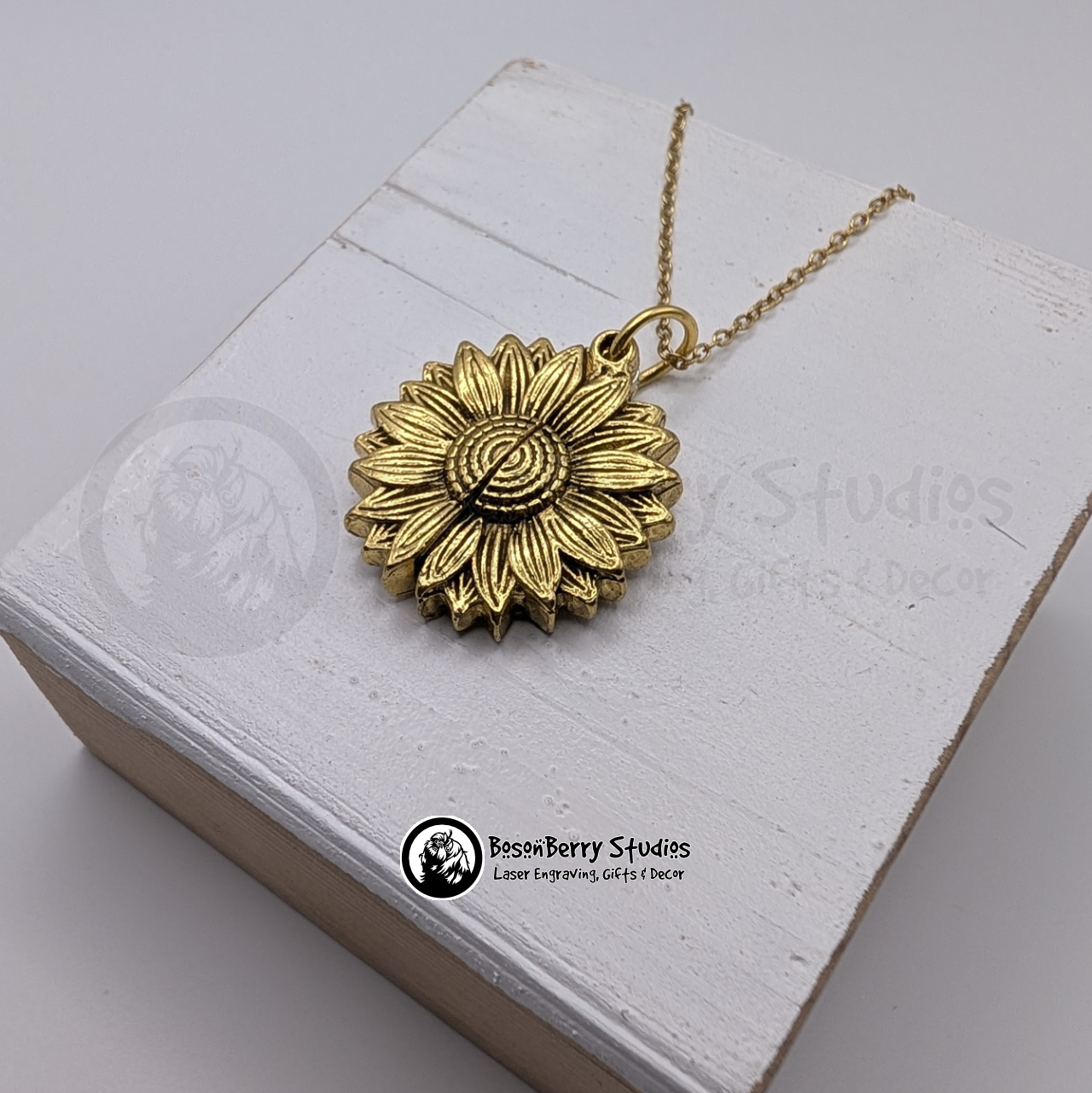 Gold sunflower pendant necklace, closed, on a white wooden stand with 'BosonBerry Studios' branding.