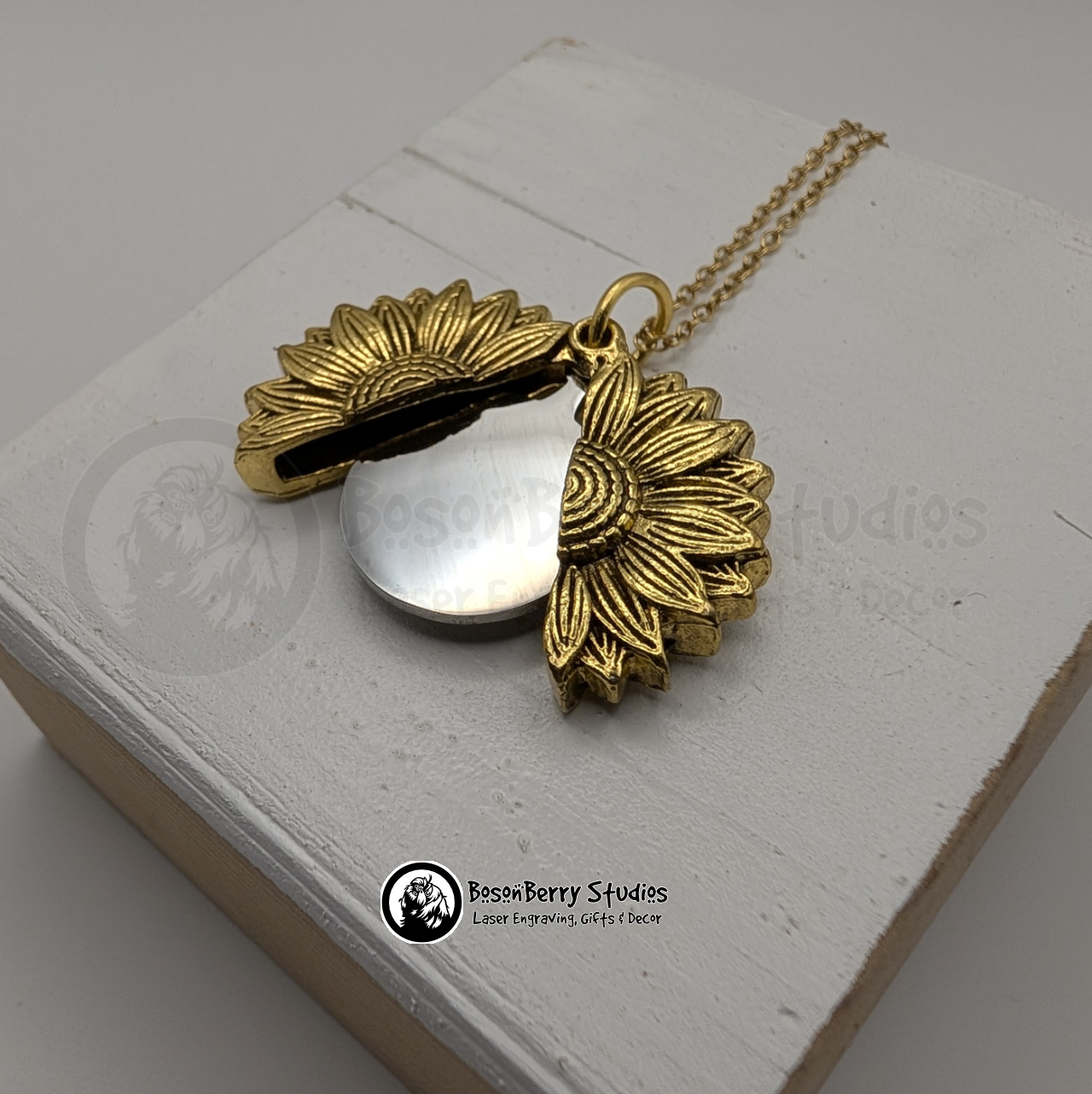 Gold sunflower pendant necklace, open to reveal a silver disc, on a white wooden stand with 'BosonBerry Studios' branding.