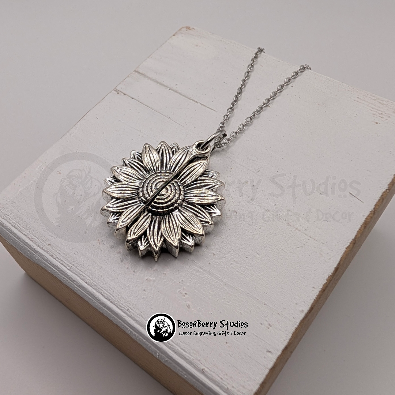 Silver sunflower pendant necklace, closed, on a white wooden stand with 'BosonBerry Studios' branding.