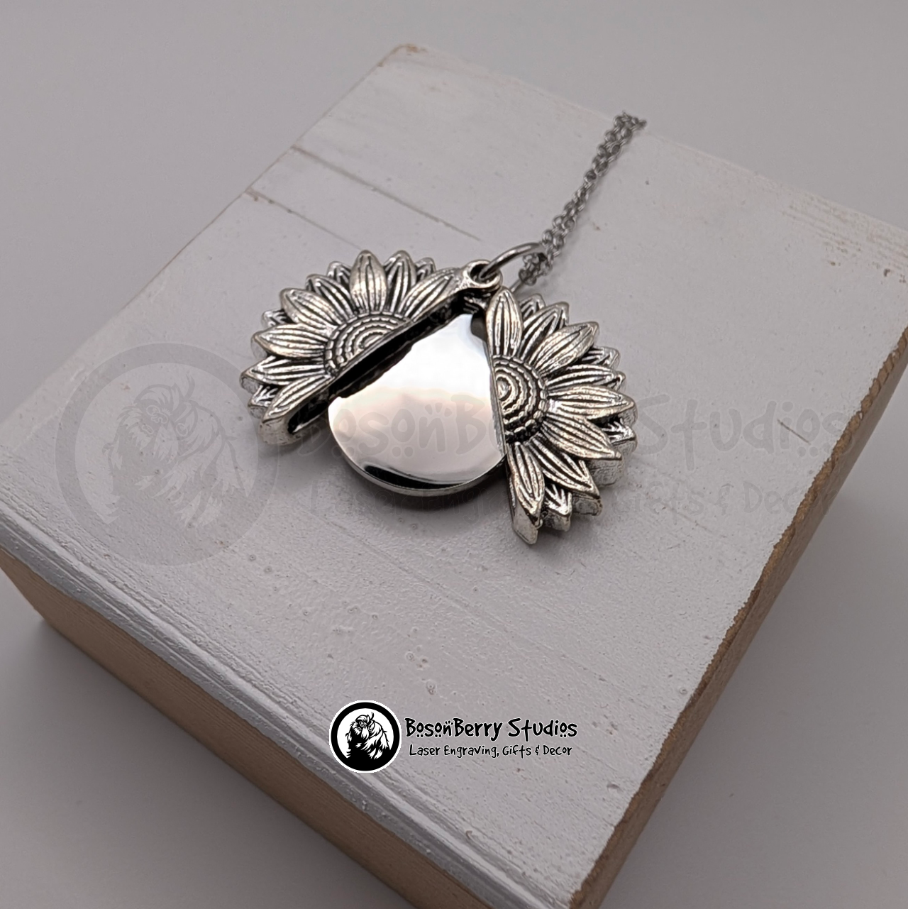 Silver sunflower pendant necklace, open to reveal a silver disc, on a white wooden stand with 'BosonBerry Studios' branding.