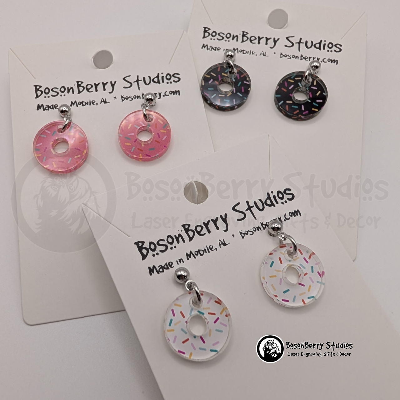 A trio of tiny donut-shaped acrylic earrings, with a colorful sprinkle pattern, one pink, one chocolate, and one white, on a BosonBerry Studios card.