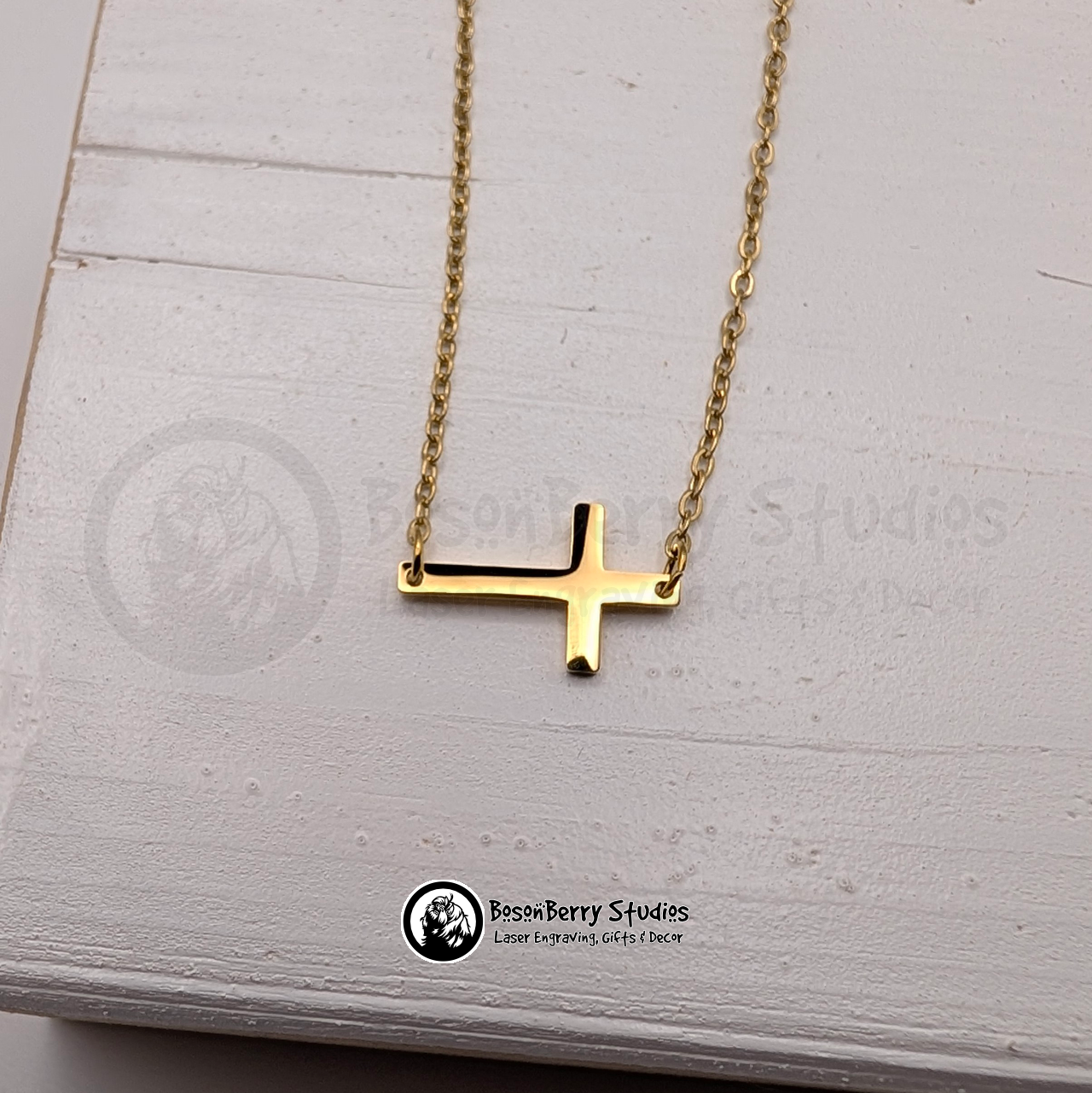 Gold necklace with a tiny cross pendant on a white wooden stand with 'BosonBerry Studios' branding.