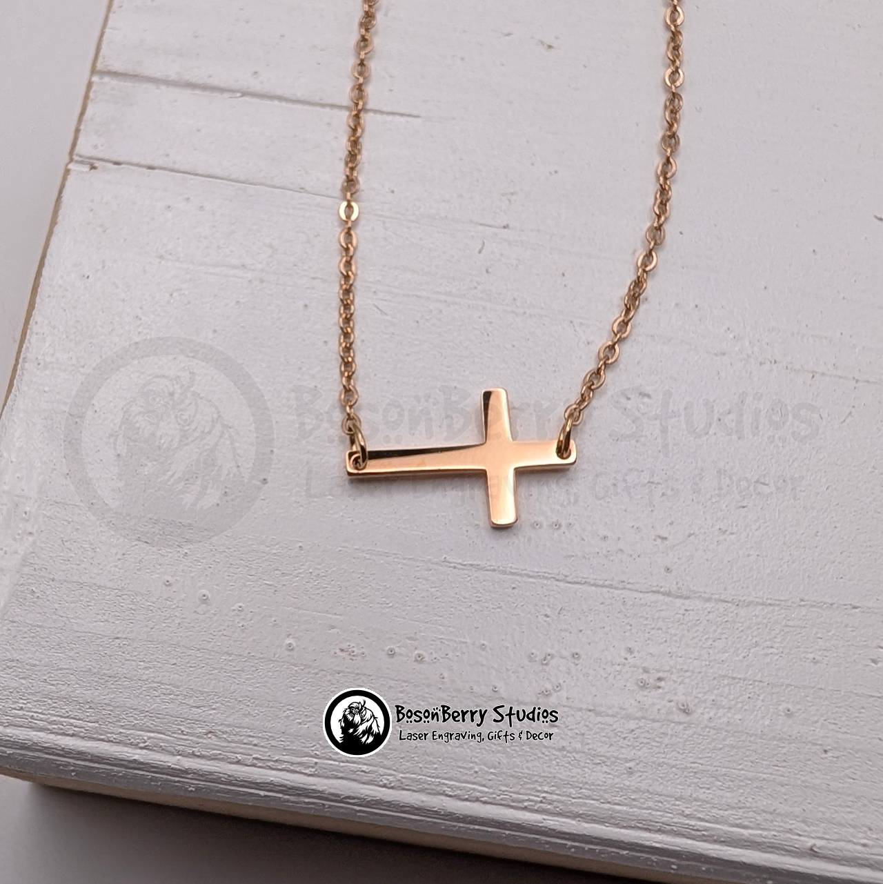 Rose gold tiny cross necklace on a white wooden stand with 'BosonBerry Studios' branding.