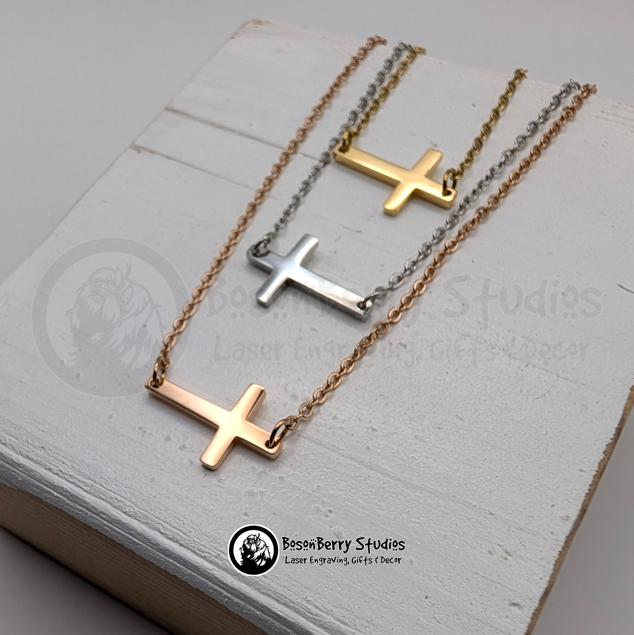 Three tiny cross necklaces, gold, silver and rose gold, on a white wooden stand with 'BosonBerry Studios' branding.