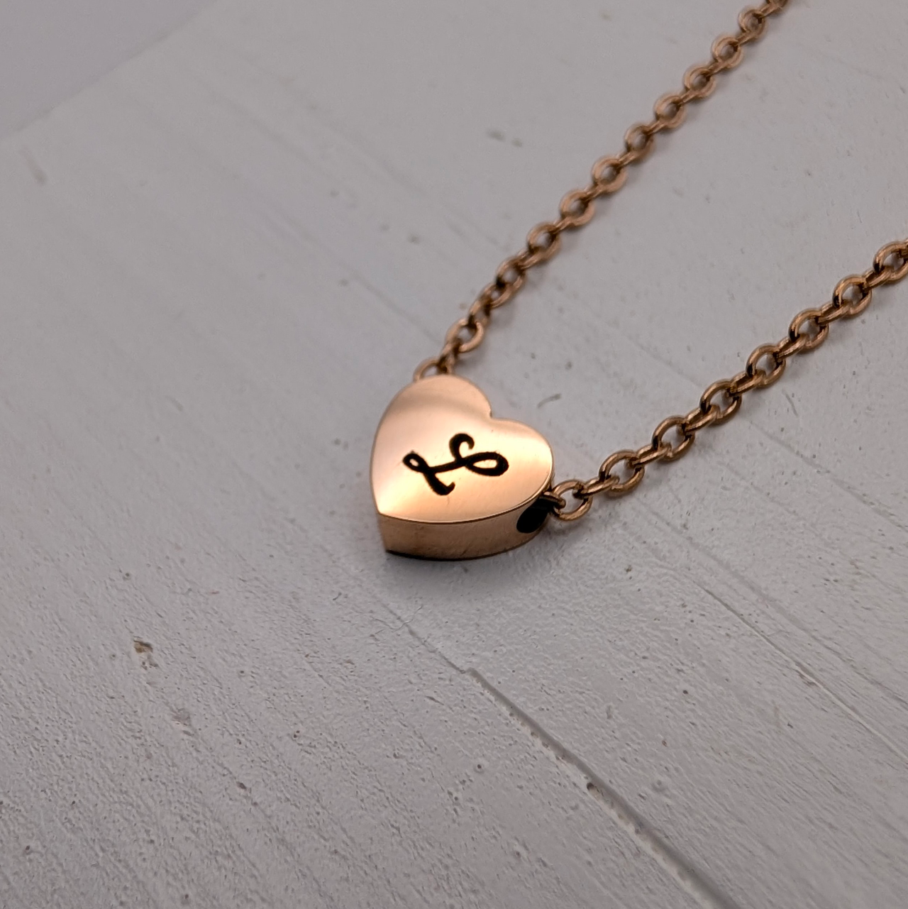Rose gold heart-shaped bead pendant necklace with a black cursive letter 'L' engraved on it, on a white wooden stand with 'BosonBerry Studios' branding.