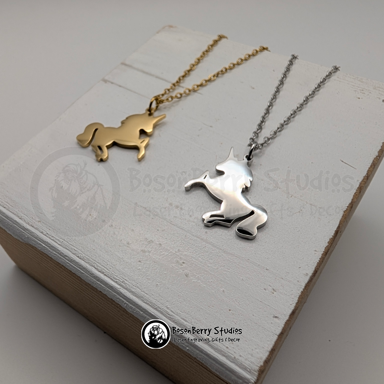 Two unicorn-shaped pendant necklaces, one gold and one silver, on a white wooden stand with 'BosonBerry Studios' branding.