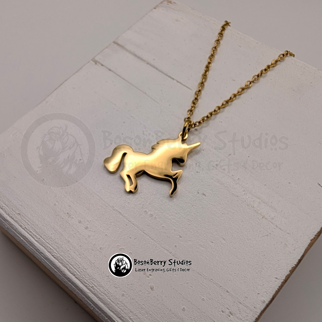 A gold necklace with a unicorn shaped pendant on a white wooden stand with 'BosonBerry Studios' branding.
