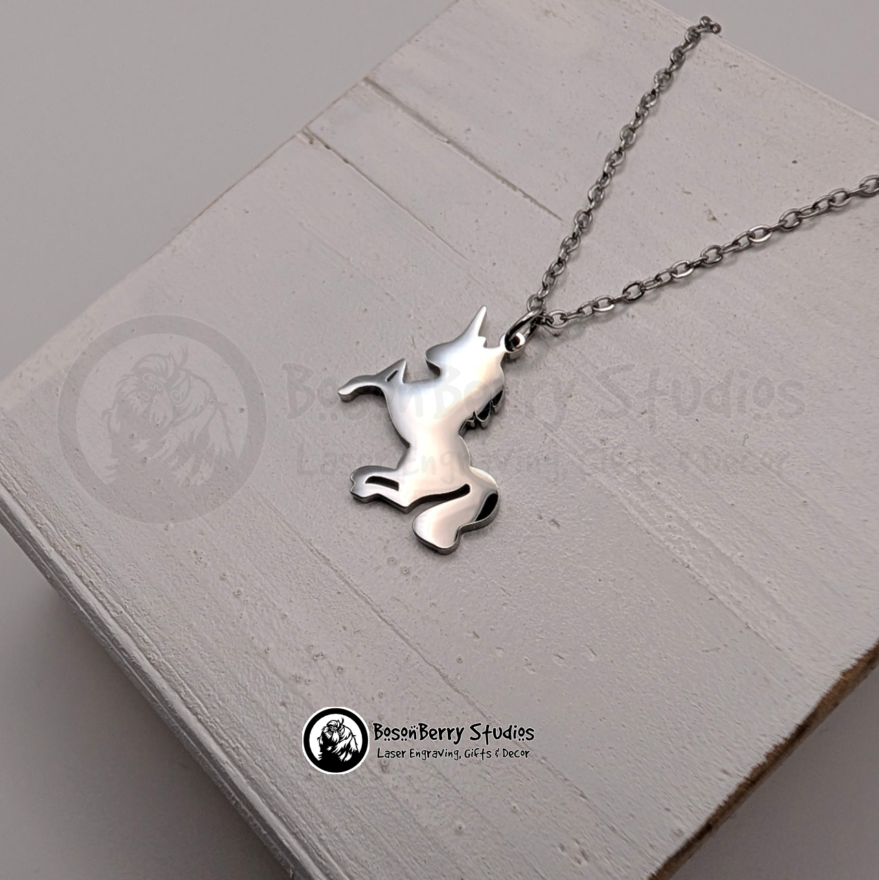 A silver necklace with a unicorn shaped pendant on a white wooden stand with 'BosonBerry Studios' branding.