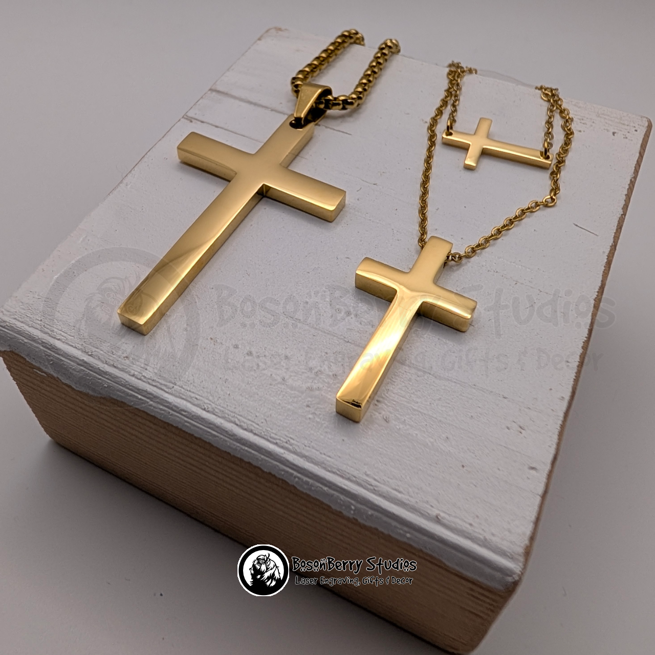 Three gold cross necklaces in tiny, regular and large sizes on a white wooden stand with 'BosonBerry Studios' branding.