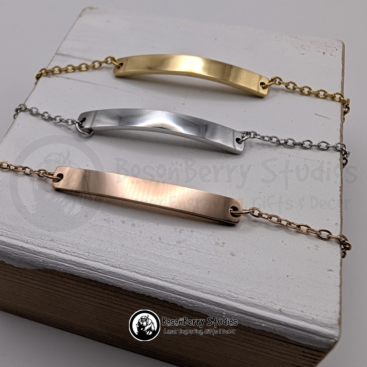 Three gold, silver, and rose gold bar bracelets on a white wooden stand with 'BosonBerry Studios' branding.