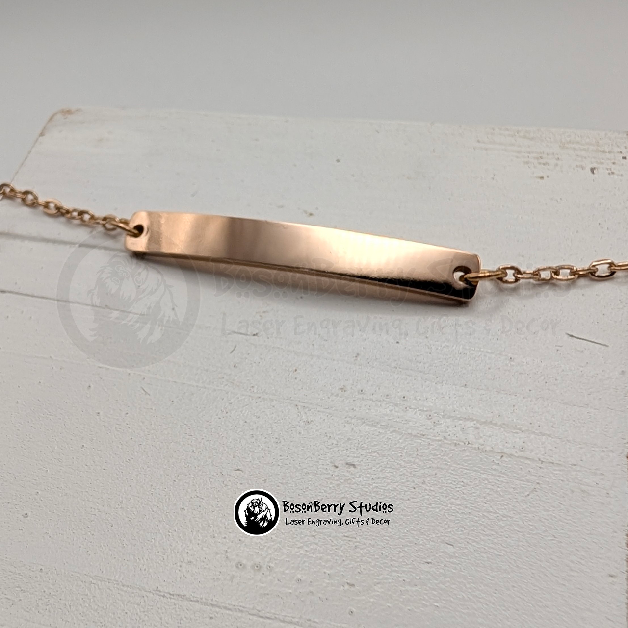 Rose gold bar bracelet on a white wooden stand with 'BosonBerry Studios' branding.